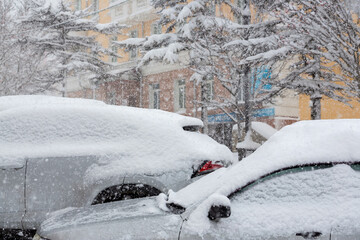 Snow-covered cars during a heavy snowfall in the city. A lot of snow on the roofs and windows of parked cars. Cold snowy winter weather.