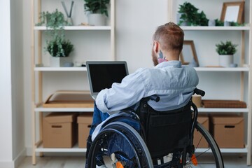 A man in a wheelchair freelancer works at a laptop at home view from the back, copy space, integration into society, health concept man with disabilities, real person