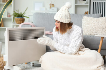 Frozen young woman in winter clothes warming near radiator at home
