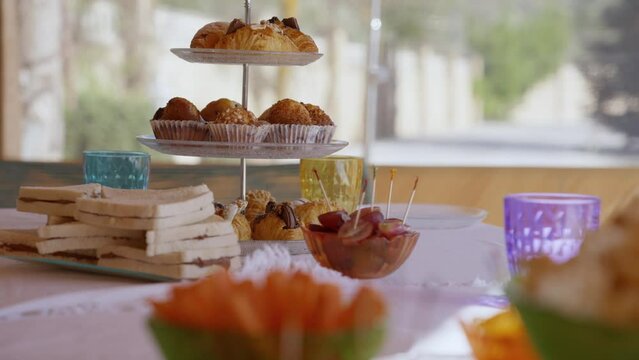 Table with sweets for a party in summer, with a tray with croissants, sandwich, crudit&eacute;s, coloured glasses and white tablecloth. Slider camera movement to left.