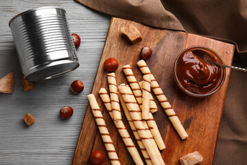 Wooden board with tasty wafer rolls, nuts, tin can and bowl of boiled condensed milk on grey table