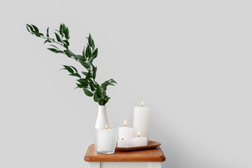 Burning candles and vase with ruscus branches on end table near grey wall