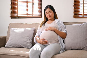 Happy Asian pregnant woman relaxes on the sofa in her living room, touching belly with love.