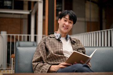 Happy Asian man in trendy outfit enjoys reading a book while chilling at the coffee shop.