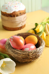 Nest with painted Easter eggs and tulip flowers on yellow table, closeup