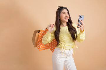 Happy smiling Asian female shopper holding a bunch of shopping bags while holding a mobile phone. Fashionable girl in yellow top and white trousers looking at camera isolated on beige color background