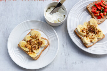 Sandwich breakfast has banana cream cheese almonds and tomato on white wood table.