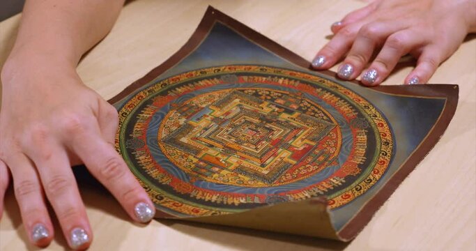 Canvas of a Nepalese tank, with a painted wheel of existence. A close-up of women's hands unrolling a scroll of a tank on a table, with an image symbolizing the wheel of existence. Wisdom of Nepal