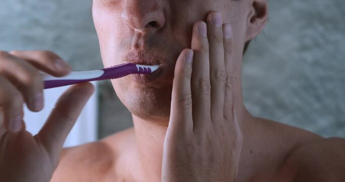 Close up young man suffers from tooth pain when brushes teeth with tooth brush, in bath. Problems with teeth, acute tooth ache, concept of caries and oral hygiene
