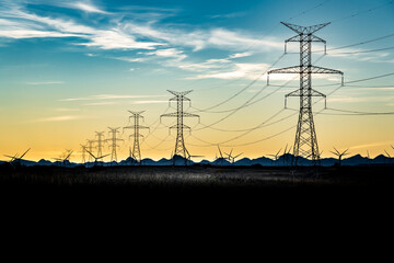 Sunset silhouette of steel lattice transmission towers and power lines overlooking distant mountains and windmills.