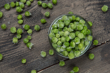 Scattered green peas on a wooden board and a glass bowl full of frozen peas with ice