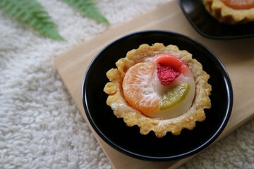Kue pia buah or Small fruit pie cake on a black round plate and wooden pad with orange amd strawberry on the top. Used to called Kue Pia Buah. White fluffy background.