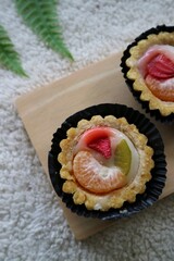 Kue pia buah or Small fruit pie cake on a black round plate and wooden pad with orange amd strawberry on the top. Used to called Kue Pia Buah. White fluffy background.