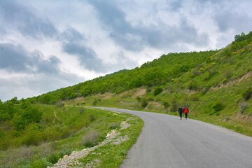 Fototapeta na wymiar Two travelers walk along an asphalt road in the green mountains