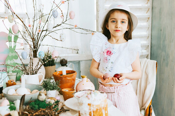 pretty little Armenian girl helps with baking for Easter on veranda on sunny spring day decorated with flowers and Easter decor, eggs, cake and willow branches, Easter family celebration