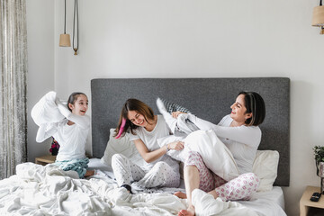 Obraz na płótnie Canvas Hispanic happy family of women, mother and daughter laughing, playing pillow fight on bed at home in Latin America
