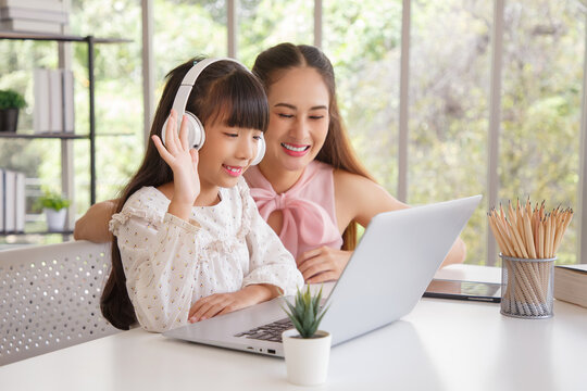 Children education and home school concept : Young asisn mother pleased to see little daughters' study online. Excited smiling child girl enjoying onlinr learning  with pleasant mom at home.