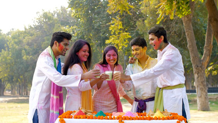 Cheerful Indian teenagers smeared in powder colors celebrating joyful Holi festival. Group of young friends wishing each other Happy Holi while having delicious Kesar Thandai at a Holi party in a p...