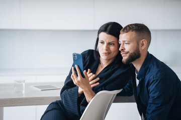 Perplexed young couple sitting at kitchen looking at phones screen with puzzled face expression upset by news. Young adult hispanic woman shows to husband, disappointed by economic crisis. Frustration