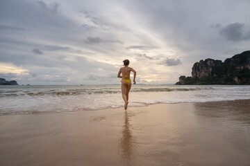 Woman wearing yellow bikini smiling and running on Railay Beach at sunset. Thailand