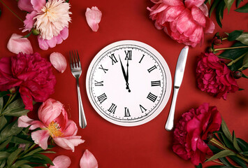Festive table setting with clock- plate, cutlery and pink peonies on red table.