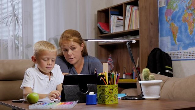 An adult mother and her schoolboy son do homework during distance learning using a tablet. The child makes an entry in a notebook. Online education and e-learning concept.