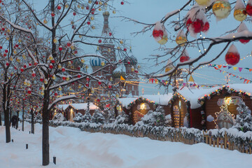 Christmas balls and glowing garlands on tree branches. Snow-covered St. Basil's Cathedral on Red Square in Moscow. Christmas market. Snowy winter cityscape.