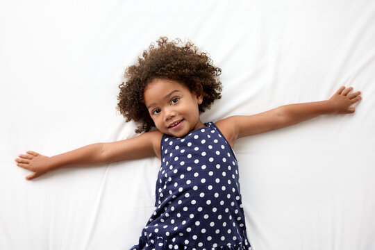 Smiling little girl with afro hair lying on white bed with arms extended