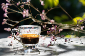 Espresso shot coffee in clear small cup with blur pink flower in background in the morning