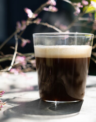 A cup of americano macchiato coffee  with smoke above and blur pink flower in the background in the morning