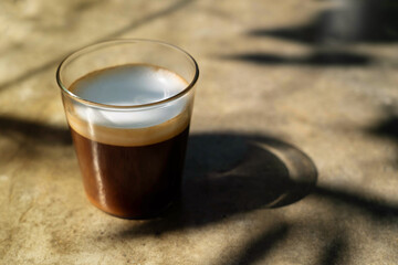 Clear cup of americano macchiato coffee  with  shadow of tree branch on desk  in the morning