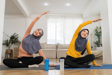 Two Asian muslim women in hijab practicing yoga on mat at home. Smiling muslim women exercising on floor in living room