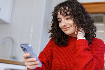 Portrait of happy Ukrainian girl browsing internet on smart phone at home. Cheerful brunette female person using modern blue mobile phone