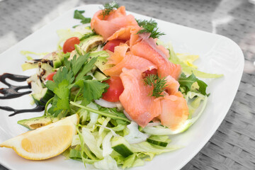 A smoked salmon and avocado salad with green lettuce, dill, cherry tomatoes and cucumber, with a slice of lemon