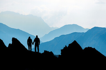 Silhouette Couple of man and woman reaching mountain top enjoying freedom and looking towards blue mountain silhouettes and sunrise. Alps, Allgaeu, Bavaria, Germany. - 567306594