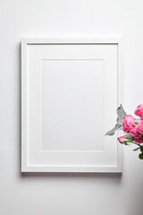 Photo frame mockup with passe-partout hanging on white wall and and bouquet of pink roses with eucalyptus. Empty blank frame and floral decor
