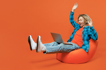 Full body overjoyed happy young IT man wear blue shirt orange t-shirt sit in bag chair hold use...
