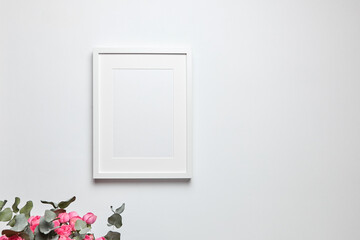 Photo frame mockup with passe-partout hanging on white wall and and bouquet of pink roses with eucalyptus. Empty blank frame and floral decor