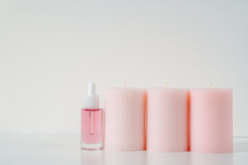 pink face oil in glass bottle with white cap and big round candles in a row. Cosmetics beauty product background