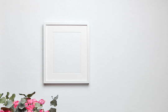 Photo frame mockup with passe-partout hanging on white wall and and bouquet of pink roses with eucalyptus. Empty blank frame and floral decor