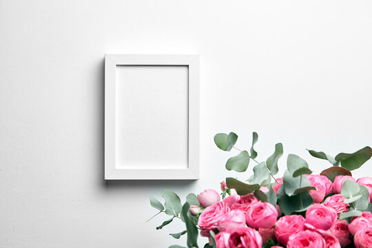 White photo frame mockup hanging on white wall and and bouquet of pink roses with eucalyptus leaves. Empty blank frame and flowers