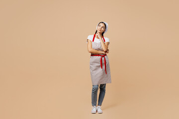 Fototapeta premium Full body minded young housewife housekeeper chef baker latin woman wear apron toque hat look aside on workspace area prop up chin isolated on plain pastel light beige background. Cook food concept.