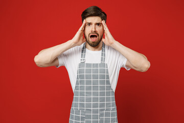 Young shocked sad disappointed dissatisfied displeased male housewife housekeeper chef cook baker man wear grey apron look camera hold head isolated on plain red color background Cooking food concept.