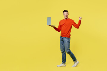 Full body side view young IT man wear orange casual clothes hold use work on laptop pc computer do winner gesture isolated on plain yellow color background studio portrait. People lifestyle concept.