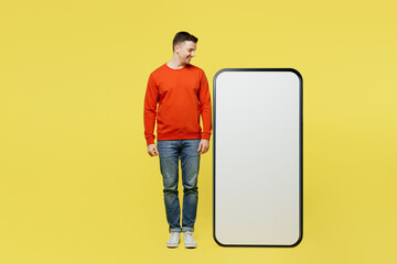 Full body smiling young man wear orange casual clothes stand near big huge blank screen mobile cell phone looking at smartphone with copy space mockup area isolated on plain yellow color background.