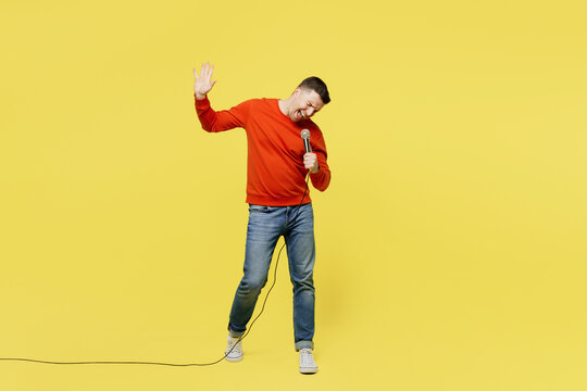Full body young expressive cool singer fun man wear orange casual clothes sing song in microphone at karaoke club isolated on plain yellow color background studio portrait. People lifestyle concept.
