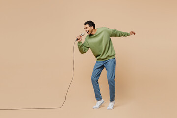 Full body expressive fun young man of African American ethnicity wear green sweatshirt sing song in microphone isolated on plain pastel light beige background studio portrait People lifestyle concept