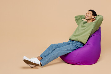 Full body young man of African American ethnicity wear green sweatshirt sit in bag chair hold hand...