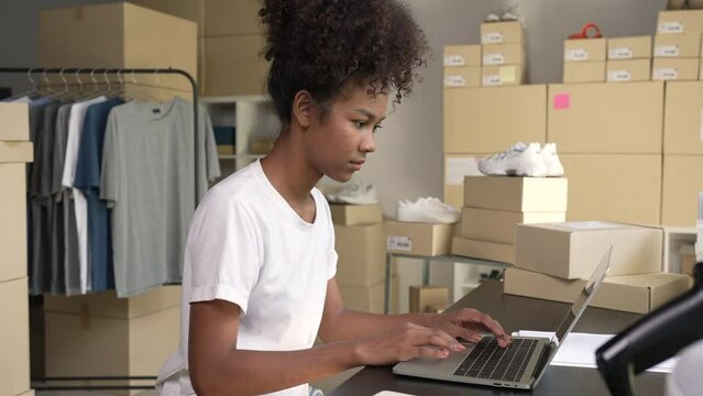 African-American woman online seller confirming orders from customer on laptop. E-commerce business owner working on laptop in store warehouse.