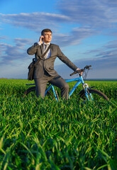 a businessman rides a bicycle on a green grassy field, dressed in a business suit, he makes a phone call, beautiful nature in spring, freelance business concept